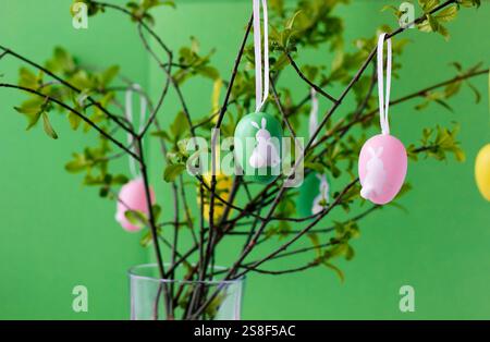 Colorful Easter eggs with cute white bunnies hanging on green tree branches in glass vase, green background. Easter concept. Easter still life. Stock Photo