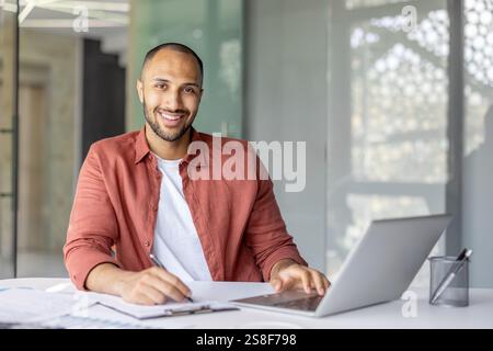 Portrait of a young successful businessman financier at his workplace inside the office, the employee is smiling and looking at the camera, writing down data on a paper report document Stock Photo