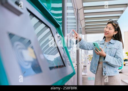 Chinese Young Woman Using Ticket Machine at Tram Stop Holding Smartphone Stock Photo