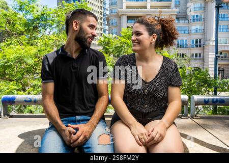 A young man and woman sit on a concrete surface in a city park, engaging in a joyful conversation with modern buildings and lush greenery in the backg Stock Photo