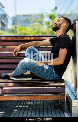 A bearded young man in casual wear enjoys a moment of laughter while sitting alone on a park bench, surrounded by greenery in a sunny urban setting Stock Photo