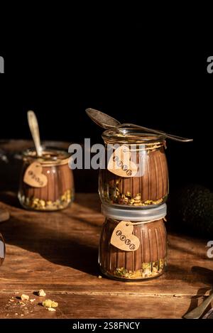Vegan chocolate avocado mousse served in glass jars, decorated with Love tags and pistachios, captured in warm sunlight on a rustic wooden backdrop. Stock Photo