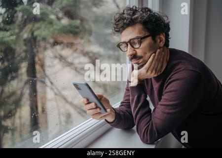 Adult middle eastern good looking man with glasses is scrolling on mobile phone in the morning standing near window. Stock Photo