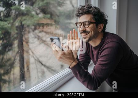 Young handsome middle eastern man with glasses having a video call on mobile phone while standing near window. Stock Photo