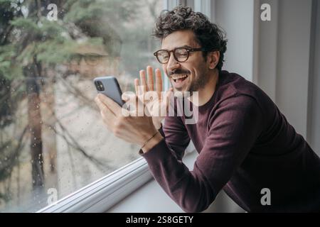 Young handsome middle eastern man with glasses having a video call on mobile phone while standing near window. Stock Photo
