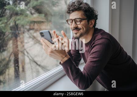Young handsome middle eastern man with glasses having a video call on mobile phone while standing near window. Stock Photo