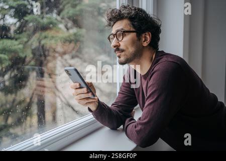Adult middle eastern good looking man with glasses is scrolling on mobile phone in the morning standing near window. Stock Photo