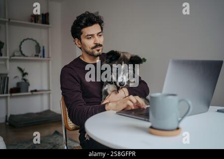 Young handsome middle eastern looking man using computer and holding cute dog in lap while working. Stock Photo
