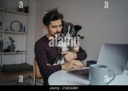 Young handsome middle eastern looking man using computer and holding cute dog in lap while working. Stock Photo
