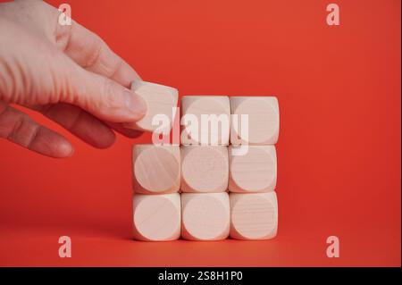 A hand places a wooden block in position on a vivid red background, creating a structure. Stock Photo