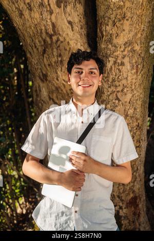 Smiling student holding laptop leaning against tree in park Stock Photo