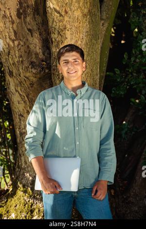 Smiling student holding laptop leaning against tree in park Stock Photo