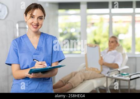 Young woman doctor posing in doctor's cabinet with patient lying on clinical chair Stock Photo
