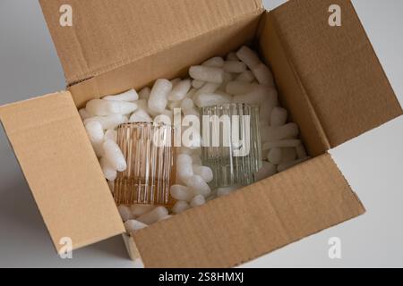 Fragile glassware in pastel shades carefully packed with protective foam peanuts inside an open cardboard box. Shipping preparation for delicate items Stock Photo