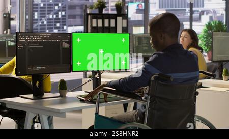 Person with disability testing and deploying programs and systems on green screen PC. Software technician in wheelchair checking code on mockup PC, sitting at desk in office, camera A Stock Photo