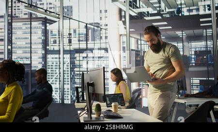 Admin at office desk utilizing cloud computing resources on laptop to train large scale machine learning models. Man monitoring company systems using notebook while standing in workspace, camera B Stock Photo