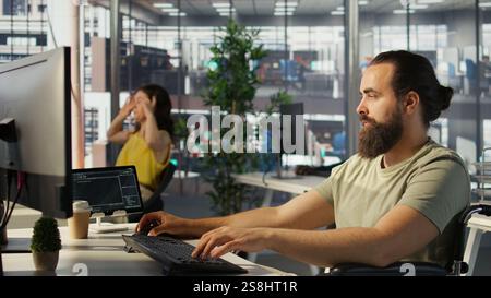 System administrator with disability in inclusive workspace training machine learning models. IT specialist with paraplegia monitoring and maintaining company systems, camera B Stock Photo