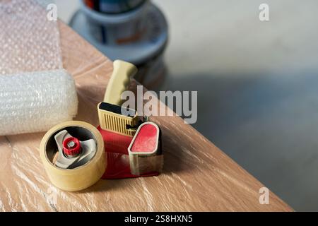 High angle of rolled transparent adhesive tape with cutter for sealing boxes on piece of furniture covered with cellophane Stock Photo