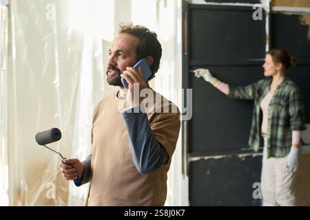 Confident young man with paintroller in hand speaking on mobile phone while standing against his wife painting wall Stock Photo