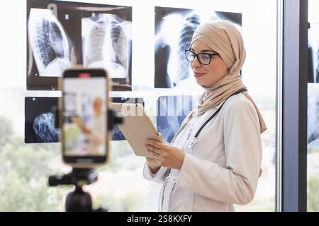 Muslim woman doctor records video lesson on phone using x-ray images. She is standing in modern office wearing hijab, smiling while explaining medical concepts. Young adult female medical professional Stock Photo