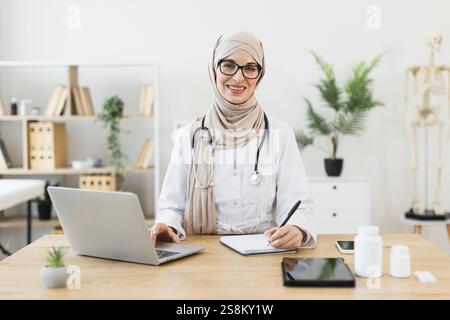 Young adult Middle Eastern woman doctor with hijab sitting at desk in medical office with laptop and notes. Stock Photo