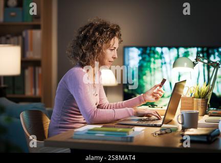 Woman sitting at the desk at home and working on her laptop, she is doing online shopping and paying using her credit card Stock Photo