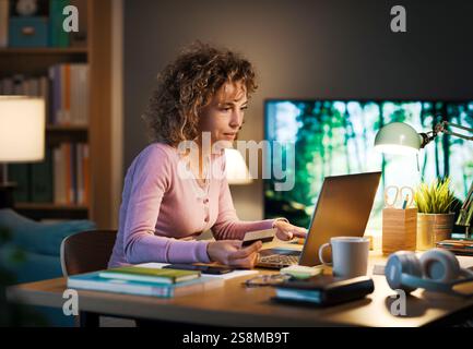 Woman sitting at the desk at home and working on her laptop, she is doing online shopping and paying using her credit card Stock Photo