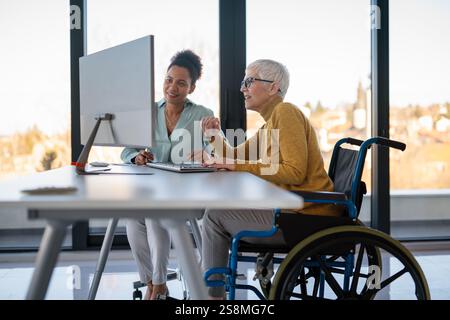 Disabled woman in wheelchair looking at computer, discussing startup project with interracial team Stock Photo