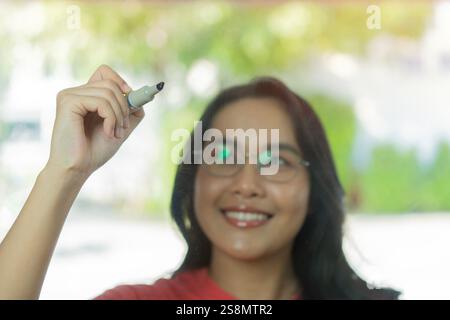 Close up image of happy young Asian woman with big pen writing on glass wall with new idea. Selective focus. Stock Photo