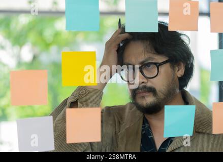 Close up view focused serious businessman writing on colorful sticky notes attached on glass wall. Business goals, daily aims, completed tasks concept Stock Photo