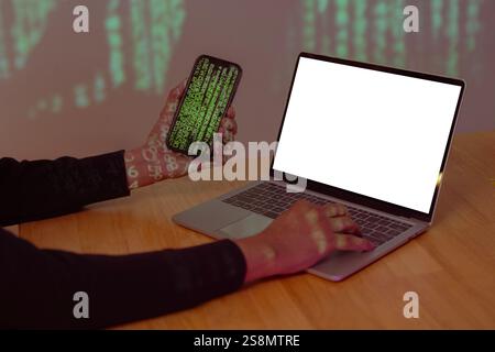 Cropped shot of hacker hands holding a mobile phone and stealing data, using laptop with blank screen. Security Concept Stock Photo