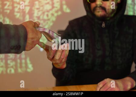 Hacker in a sunglasses with laptop receive money from employers secretly over green binary code background. Selective focus on hands. Stock Photo