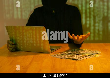 Hacker wearing black hooded with laptop hacking and stealing big data and finance information showing hand over money with green binary code backgroun Stock Photo