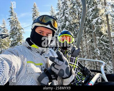 Two friends wearing ski gear riding a chairlift through a snowy mountain forest on a bright winter day. Stock Photo