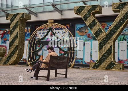 Young man browsing phone and smoking a cigarette sat on a public bench in front of large Christmas decoration of the word Joy. Exeter, Devon, UK Stock Photo