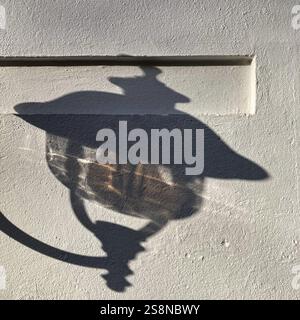 Shadow of an ornate vintage street lamp cast sharply on a white textured wall under sunlight. Stock Photo