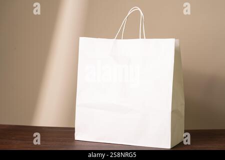 A plain white paper shopping bag with handles, placed on a wooden surface against a neutral background. Stock Photo