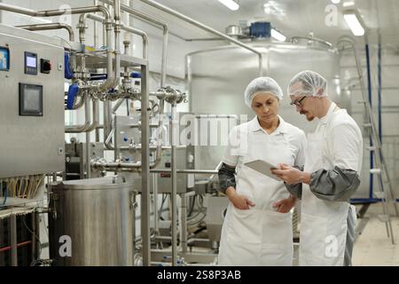 Two factory workers standing in large industrial facility, wearing white uniforms and hairnets, looking at a tablet. Machinery and stainless steel tanks in background Stock Photo