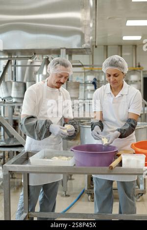 Two professionals in protective clothing and hairnets standing side by side in a commercial kitchen preparing food using various utensils and equipment Stock Photo