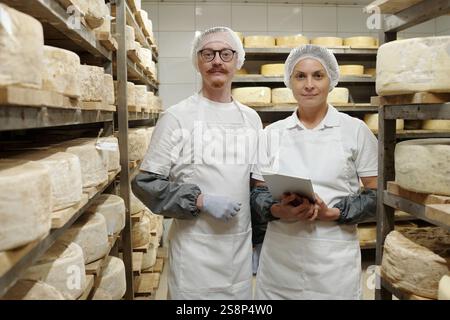 Two dairy workers wearing white uniforms and hairnets examining cheesewheels in warehouse. Shelves filled with aging cheese surround them, showcasing meticulous dairy production Stock Photo