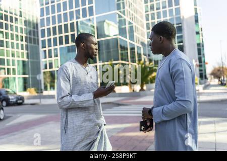 Two senegalese businessmen wearing traditional dashiki attire are discussing business outdoors, with one holding a smartphone Stock Photo