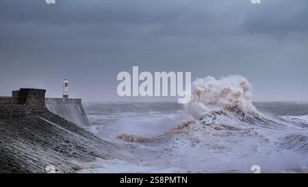 A huge winter storm hitting the coast of the UK. Giant waves crash into a breakwater, with a lighthouse. The sky is grey and moody Stock Photo