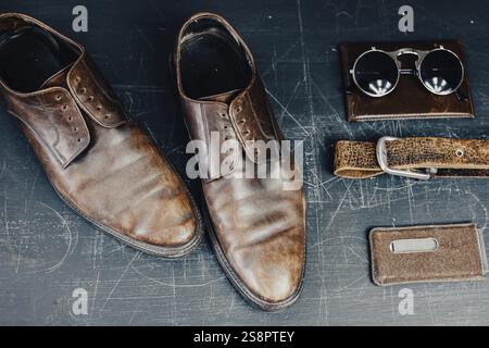 Brown leather shoes with sunglasses, belt, and wallet on a scratched dark surface Stock Photo