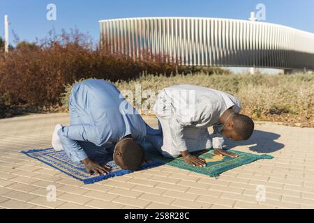 Two senegalese men wearing traditional dashiki clothing are kneeling on prayer rugs, performing salat during the holy month of ramadan Stock Photo