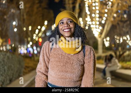 Portrait of a young black woman wearing winter clothes, smiling in a christmas decorated city at night Stock Photo