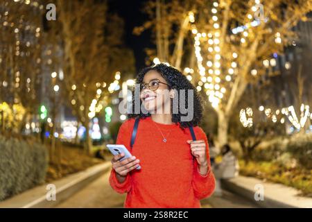 Smiling student walking in a decorated city using her smartphone during christmas holidays Stock Photo