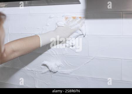 Gloved hand spreading foam cleaner on white tiled wall Stock Photo
