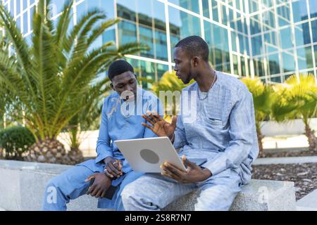Two senegalese businessmen wearing traditional dashiki attire are having a discussion while using a laptop outside a modern office building Stock Photo