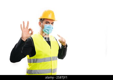 Adult male builder pointing index finger at medical or surgical disposable mask making okay gesture using fingers with blank copyspace isolated on whi Stock Photo