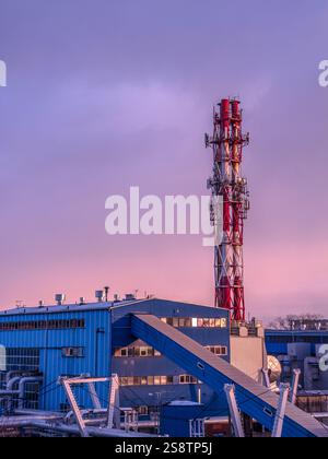 A large industrial plant featuring a prominent smokestack, surrounded by buildings and pipelines, photographed during sunset with a captivating magent Stock Photo